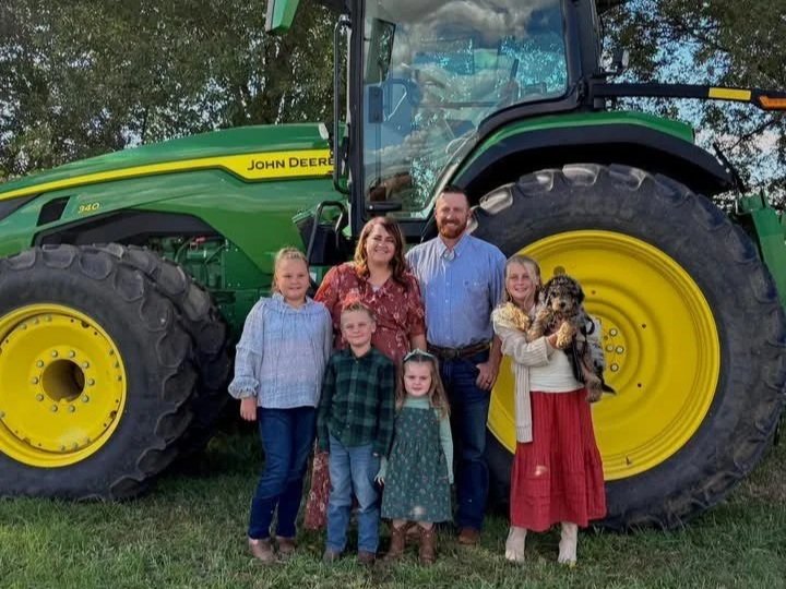 Family portrait in front of a John Deere tractor, featuring two adults and four children of varying ages, all smiling together. The scene is set outdoors with greenery in the background, showcasing a rural lifestyle.
