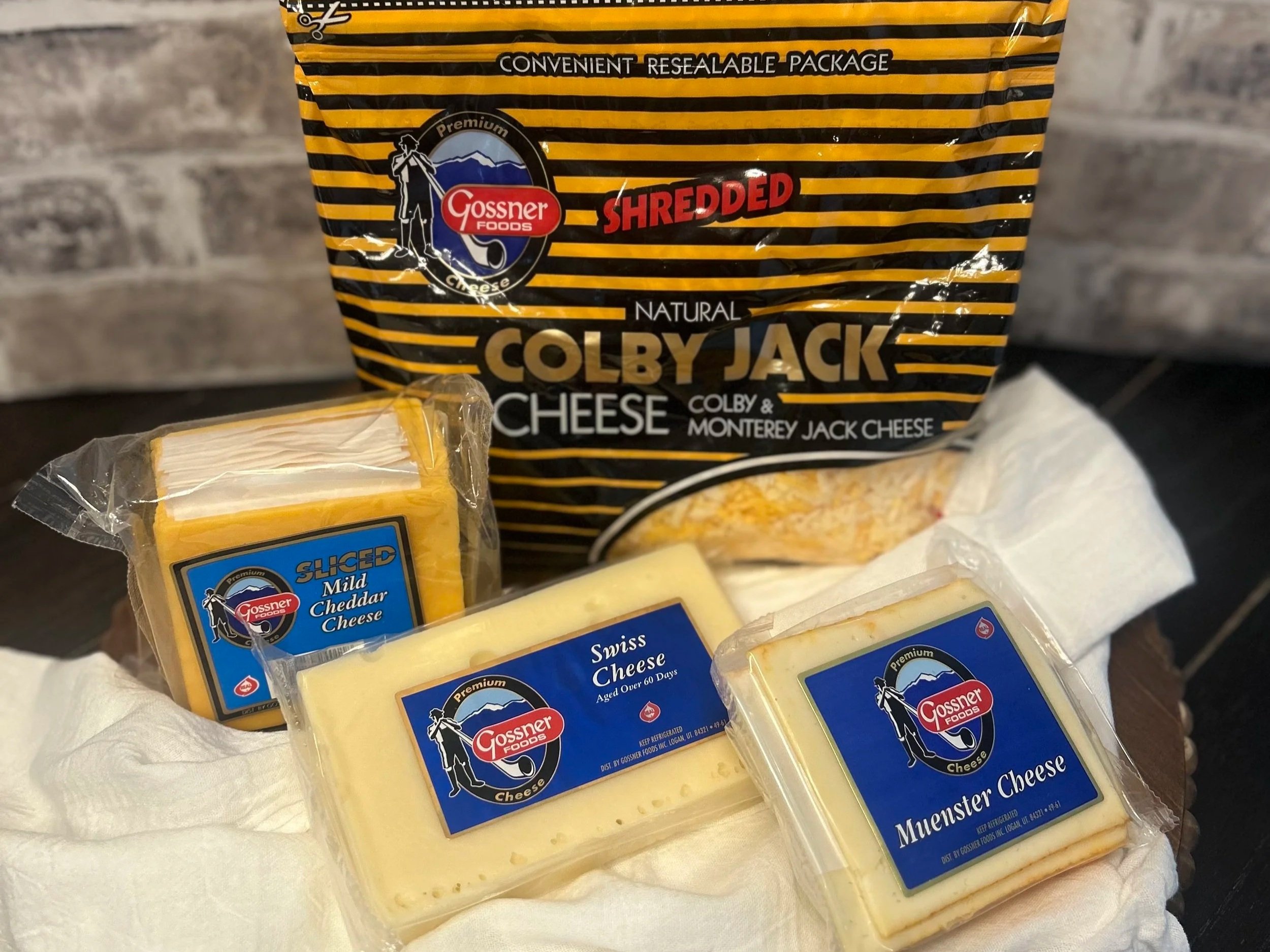 Gossner Foods cheese assortment featuring shredded Colby Jack cheese in a resealable package, along with slices of mild cheddar cheese and blocks of Swiss and Muenster cheese on a white cloth backdrop.